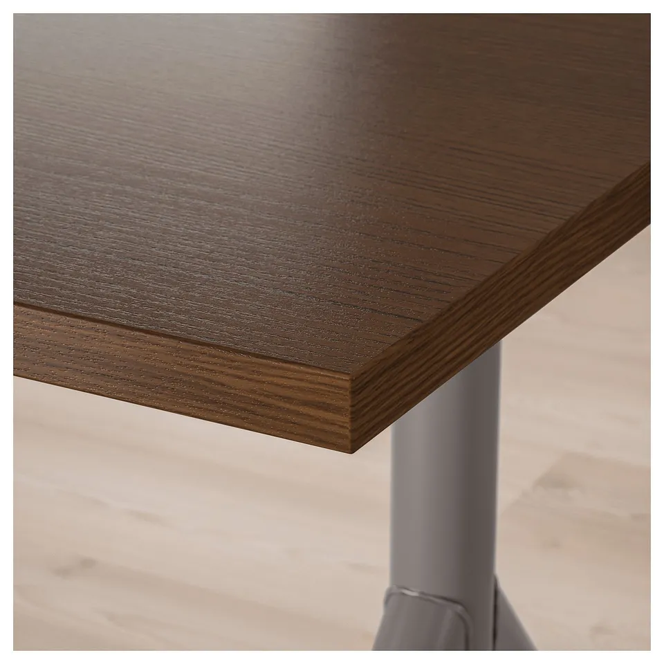 Close-up of IDÅSEN desk top showing dark wood grain pattern, smooth finish, and sturdy metal leg support.
