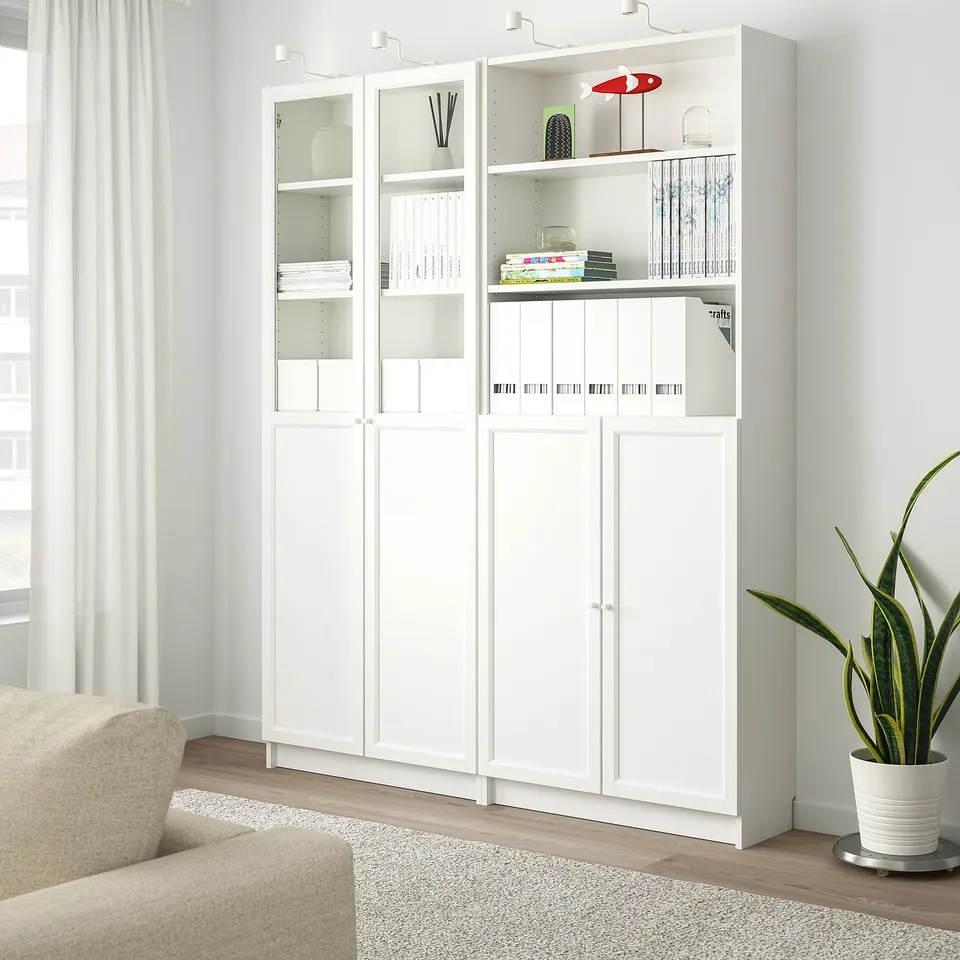 White BILLY bookcase with glass doors, holding books and decorations. Placed near a window, with a beige armchair and a snake plant.