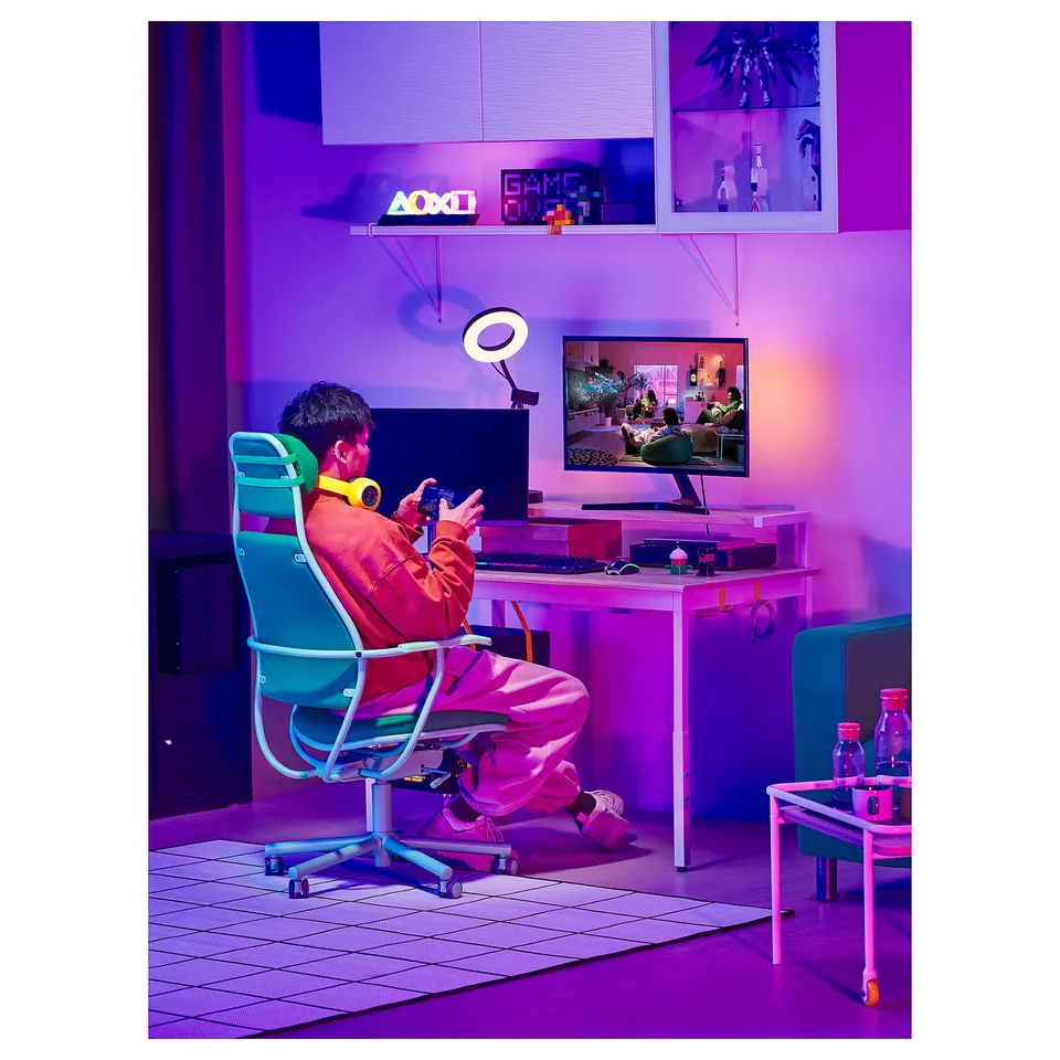 Neon-lit gamer in red hoodie on turquoise chair, gaming setup with monitor showing a game.