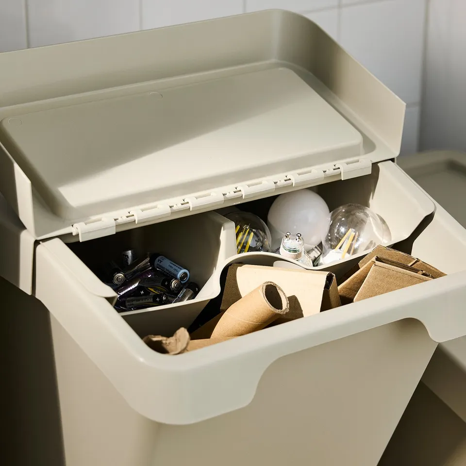 Open white SORTERA bin with lid up, showing stored items: batteries, light bulbs, cardboard, and pipes.