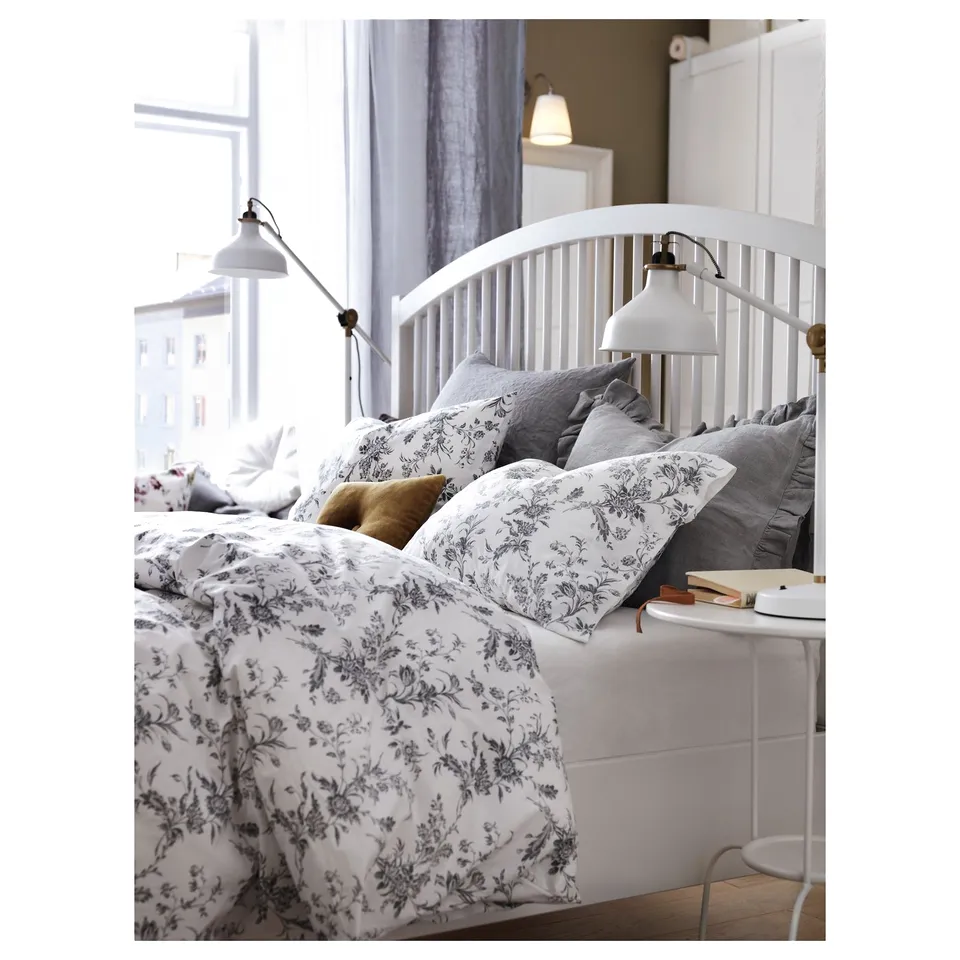 Modern white wooden bed, with floral bedding and grey pillows, beside a window.