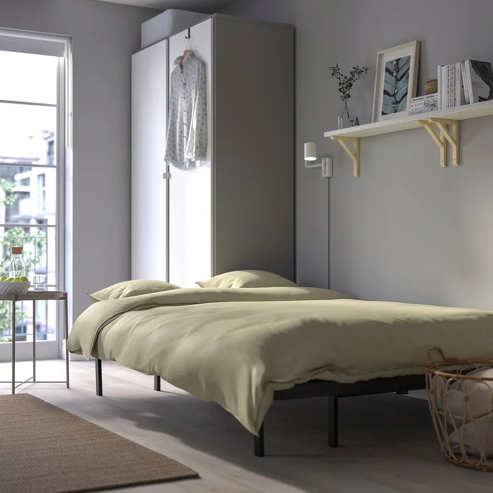 A minimalist bedroom with a double bed featuring light beige bedding, positioned against a wall with mounted shelves holding books and a vase.