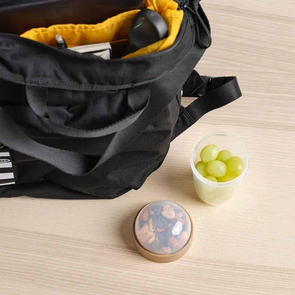 Black tote bag open on a wooden table, revealing snacks inside. Next to it, grapes in a plastic cup and a circular container with mixed nuts.