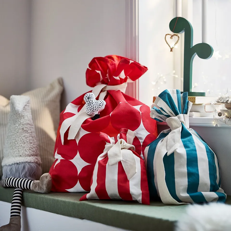 Three gift bags: two red/white, one turquoise, with ribbons. Santa & cushions on green surface. Festive decor.