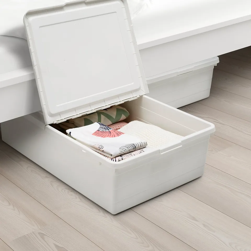 Two white plastic storage boxes under a bed, one open revealing folded blankets and a book inside.