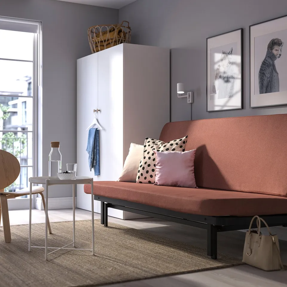 Modern living room with NYHAMN sofa-bed in terracotta, under a tall wardrobe, lit by a large window.