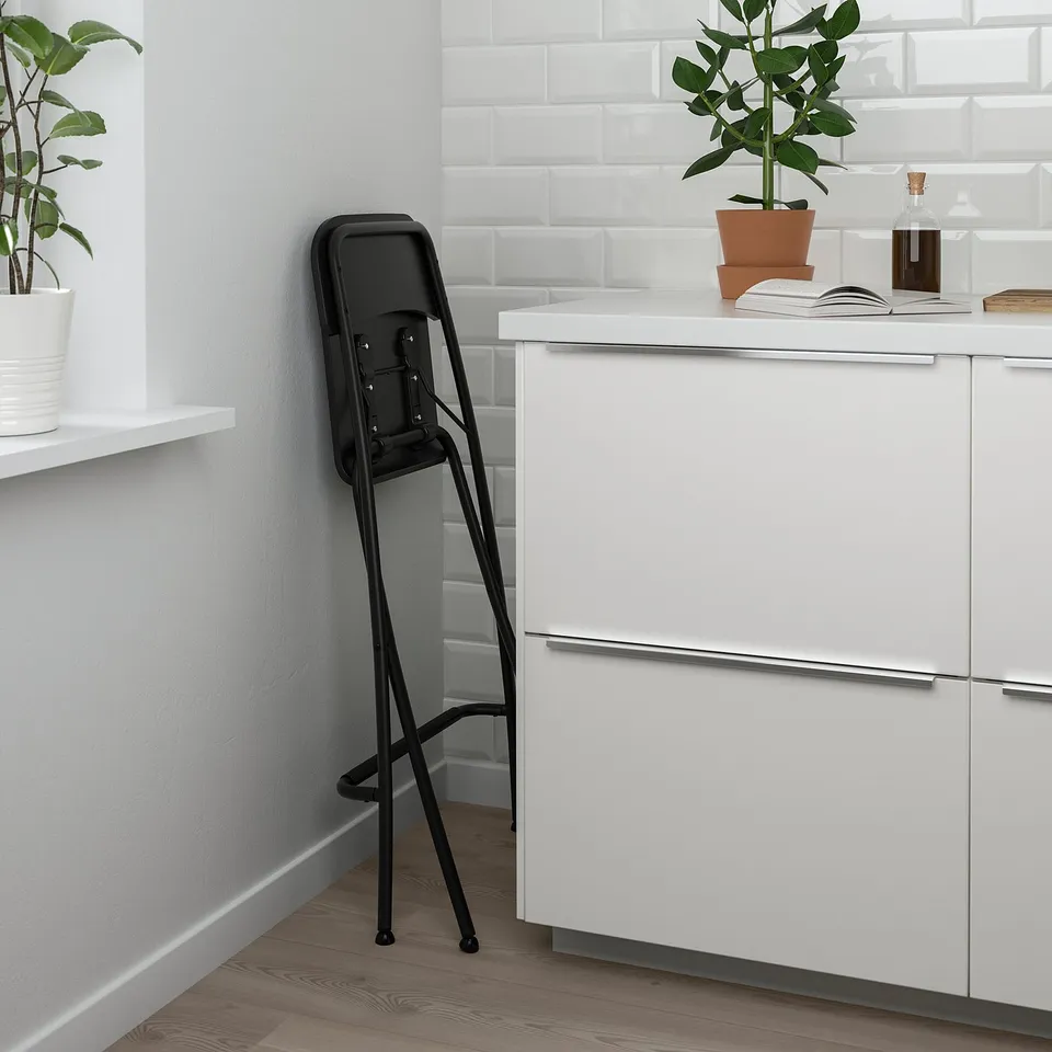 Black bar stool folded against white cabinet. Has built-in safety lock.