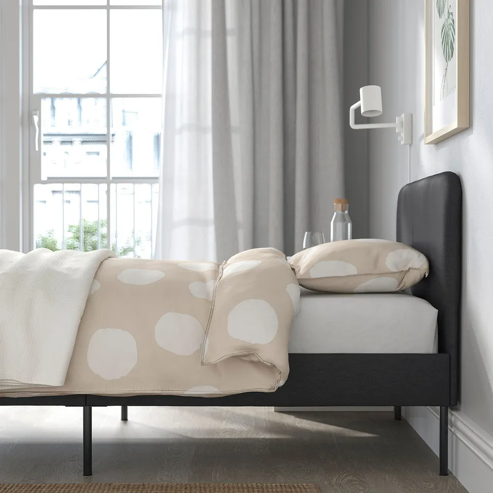 Modern bedroom showcasing the SLATTUM bed frame with soft upholstery, a padded headboard, and natural light streaming through a large window.