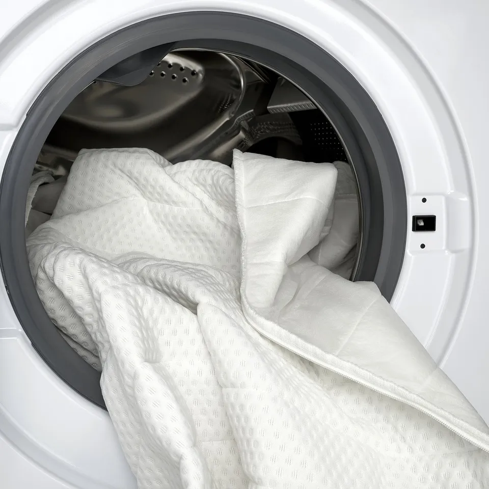 Image shows a white mattress pad inside a washing machine, ready for cleaning. The pad high-resilience foam providing a firmer sleep surface.