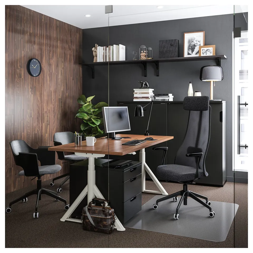 Modern office setup with black chairs, wooden desk, and storage unit. Includes a computer monitor, lamp, and plants.