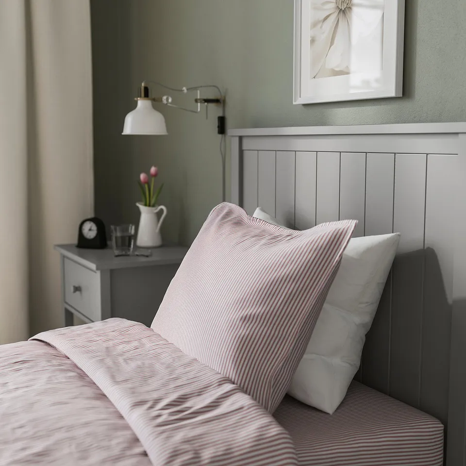 Cosy bed with striped pillows and wooden headboard, beside table with lamp and flowers.