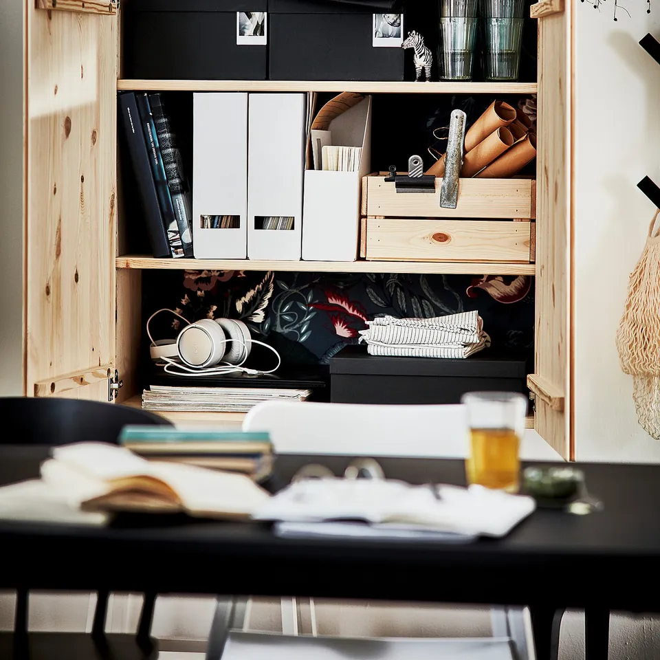 Open wooden cabinet displaying office supplies, headphones, and books. Light wood colour, square shape, multiple shelves.