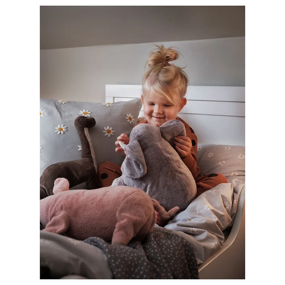 A young child sits on a bed holding a grey stuffed elephant cuddly toy. The bed has a beige comforter with white daisy designs.