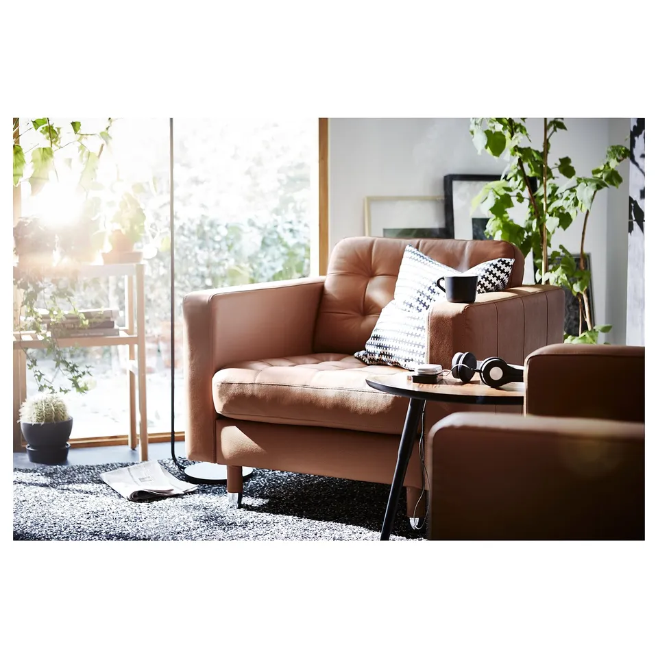 Modern living room with tan leather armchairs, geometric throw pillows, wooden side table, black rug, and large window with plants.