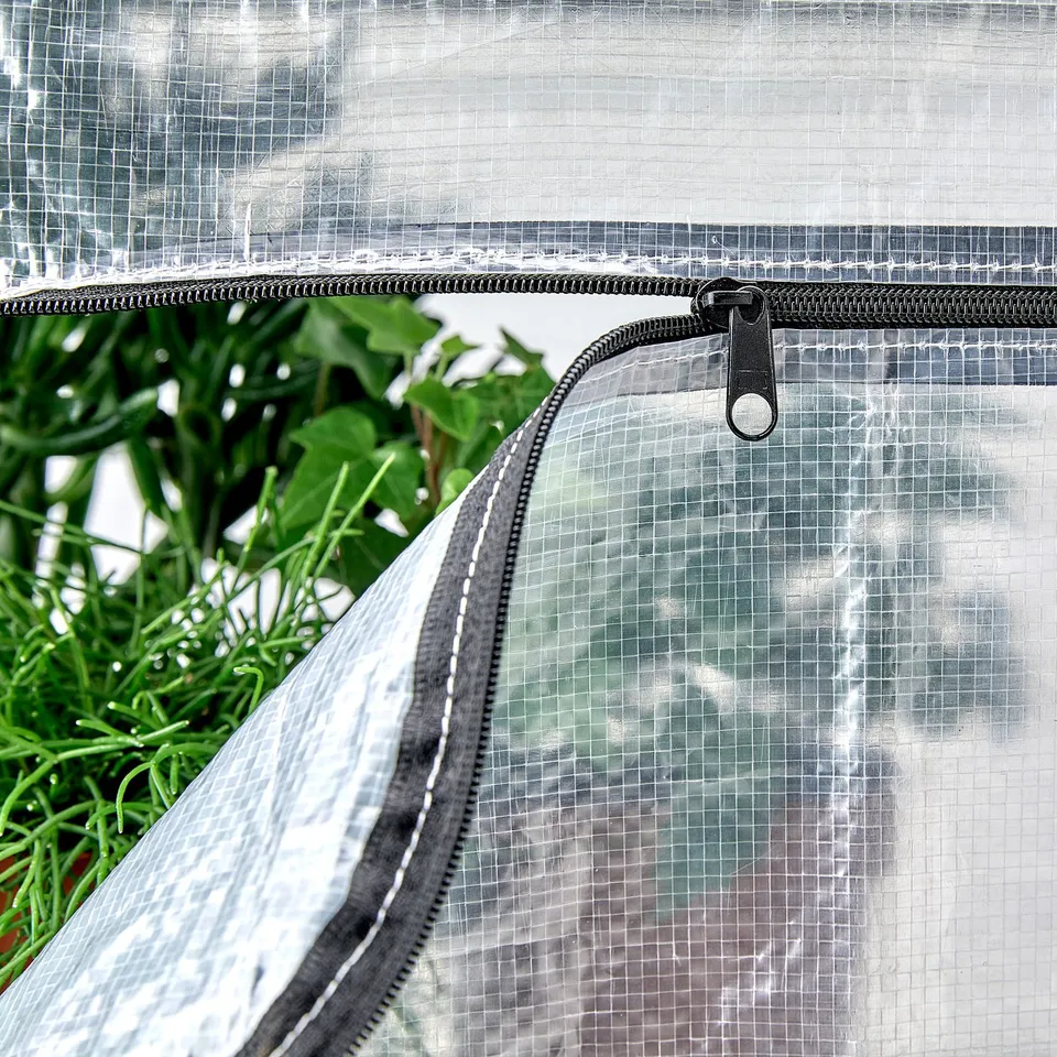 Clear plastic greenhouse cover with zip, protecting green plants; sturdy and well-made.