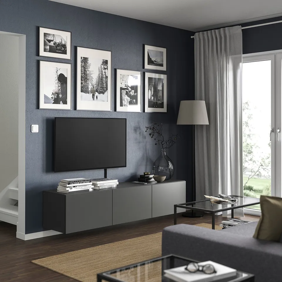 Modern living room with grey BESTÅ tv bench, flat screen tv, framed photos, lamp, books, and glass coffee table. Large windows with curtains.