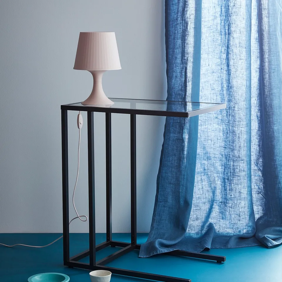 Minimalist table lamp on glass stand, with blue curtain background.