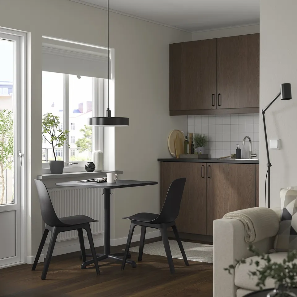 Modern kitchen with black chairs, wooden cabinets, and a table under a hanging lamp, near a white couch and large window.