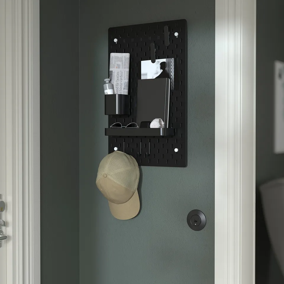 A wall-mounted pegboard organiser with shelves and hooks holding items like a cap, bottles, and papers, part of the SKÅDIS system.