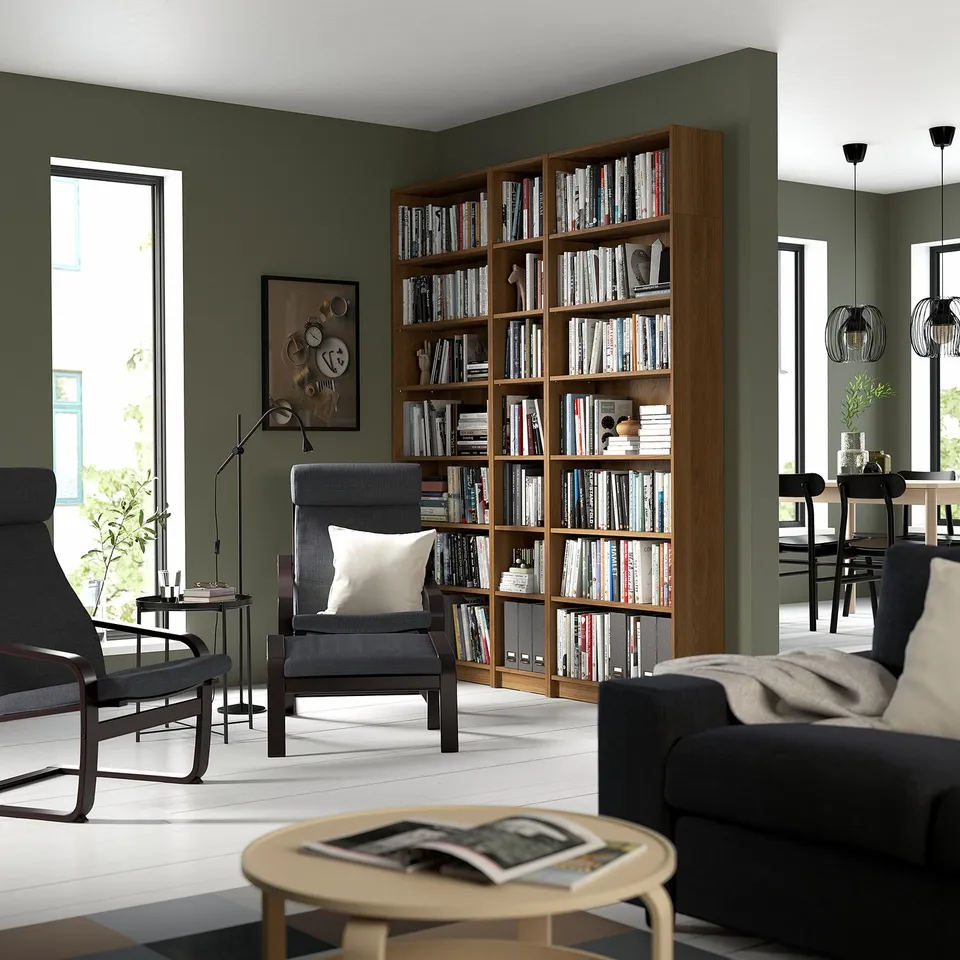 Modern living room with tall BILLY bookcase, filled with books, grey chairs, coffee table with magazines, and dining area. Green walls, white floors.