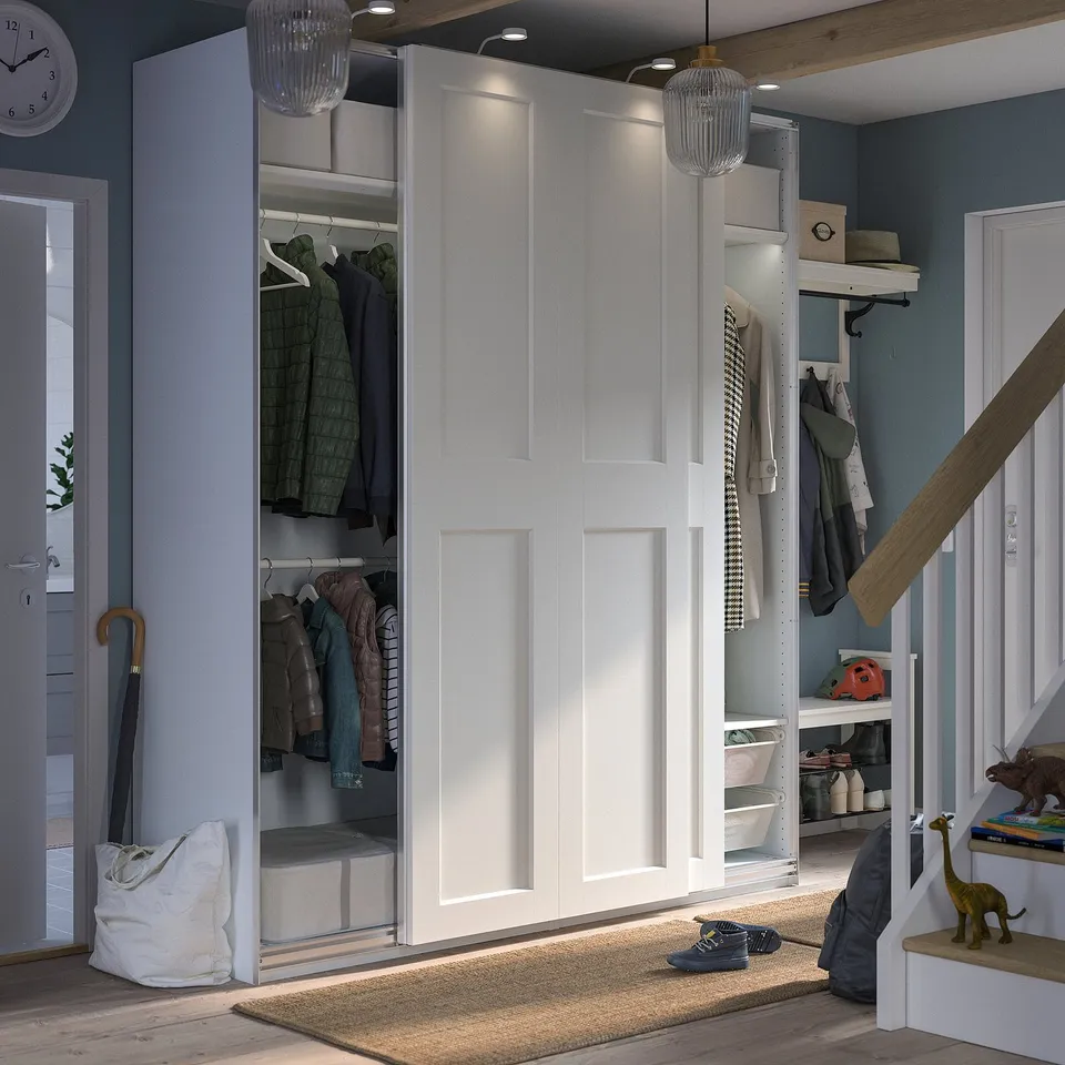 Modern white PAX closet with sliding doors reveals organised clothing and accessories. Integrated lighting and mesh basket for efficient storage.