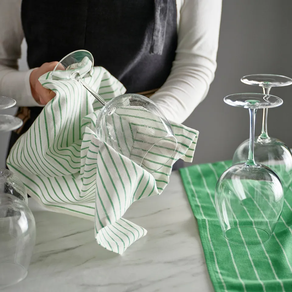 Person dries wine glasses with green-striped cloth. Glasses stand on green-striped napkin on marble table.