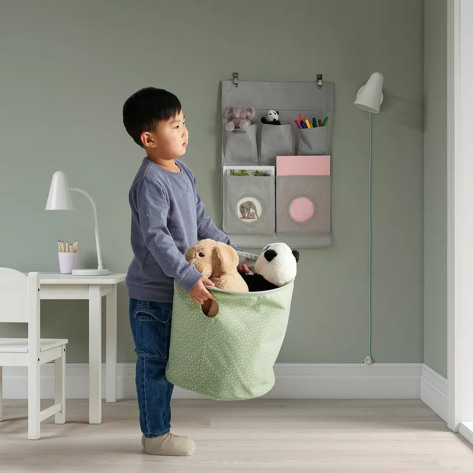 A child holding a green fabrics bag with stuffed animals. The wall behind has a hanging organiser with pockets and hooks visible.