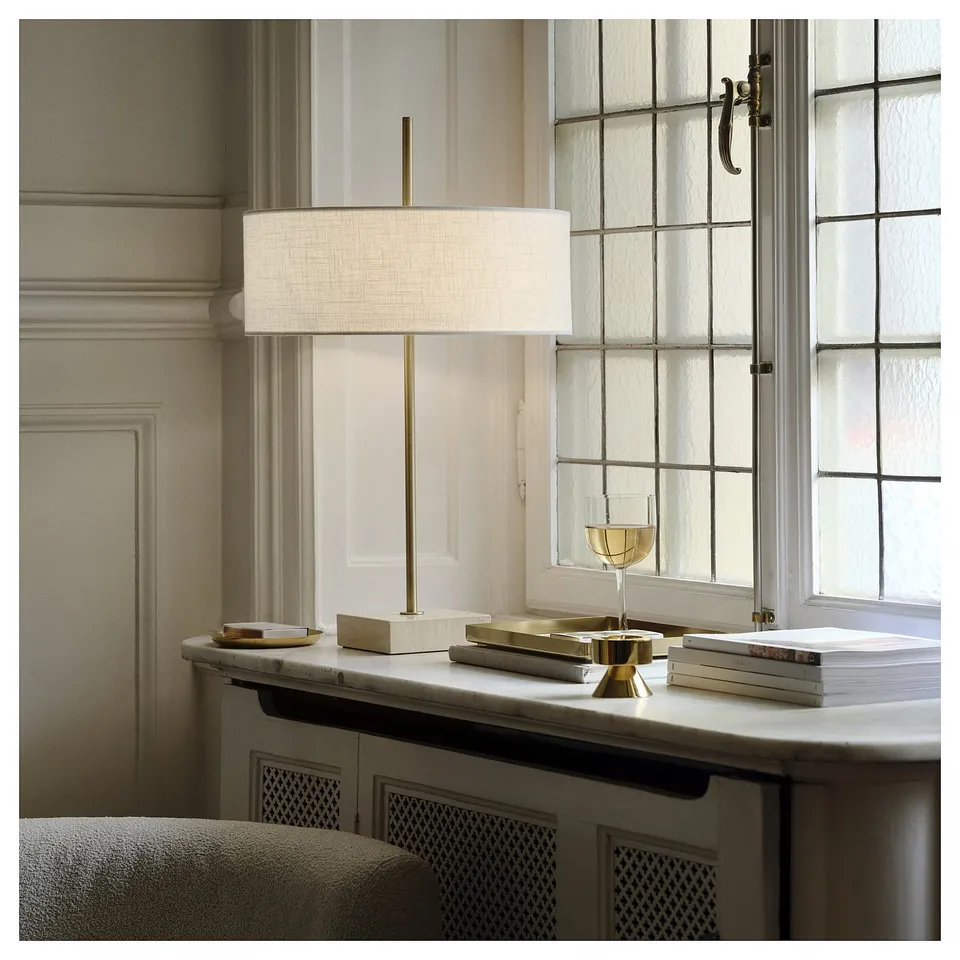 Modern lamp with white shade on travertine base near window, illuminating cosy nook with books and golden glass.
