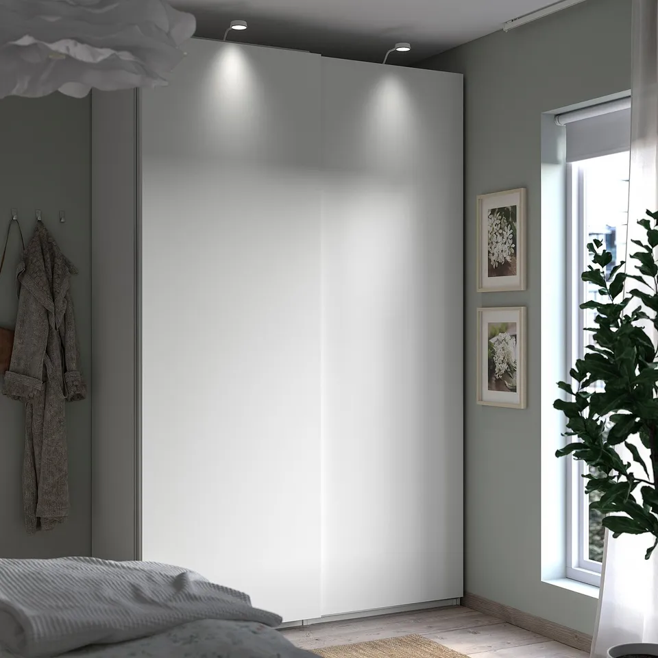 Modern bedroom with white PAX wardrobe, wall art, and green plant by window.