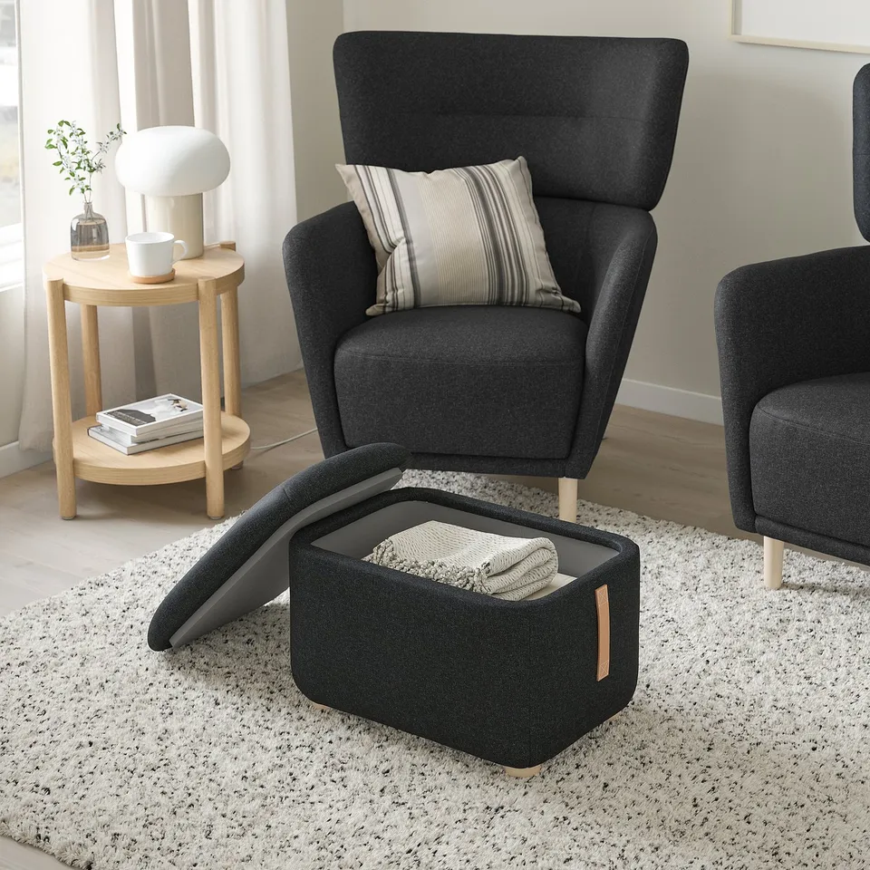 Grey wing chair with footstool, striped pillow, beige side table with lamp.