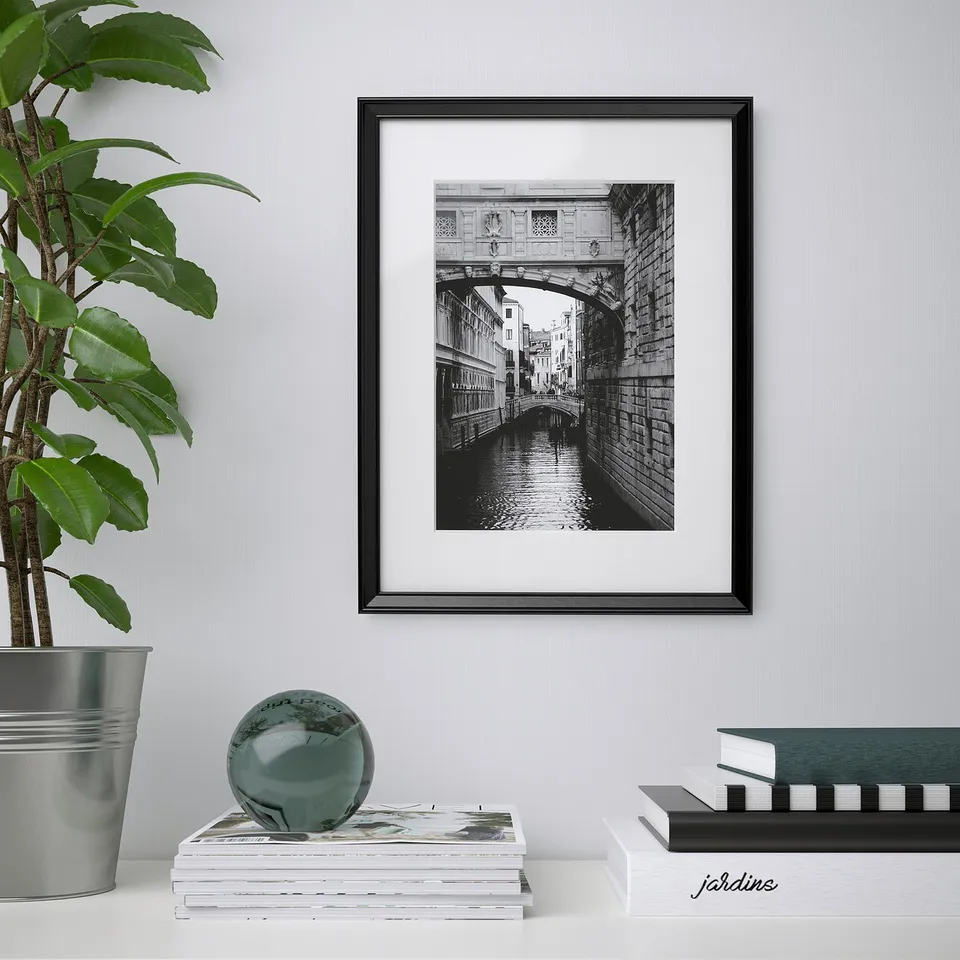 Framed black-and-white bridge over canal hangs on wall above stacked books and sculpture. Plant left.