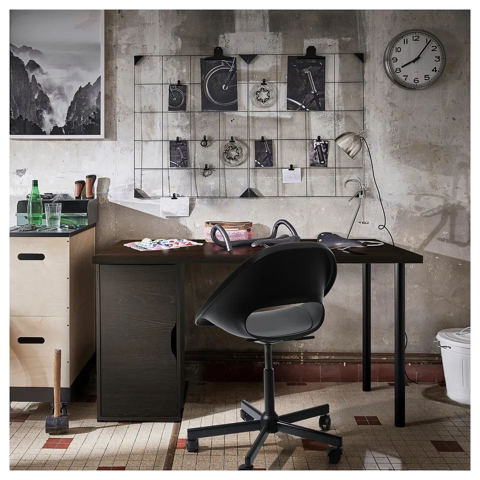 Office space with desk, chair, and wall clock. Desk has wooden top and storage. Wall has grid and photos. Tiled floor.