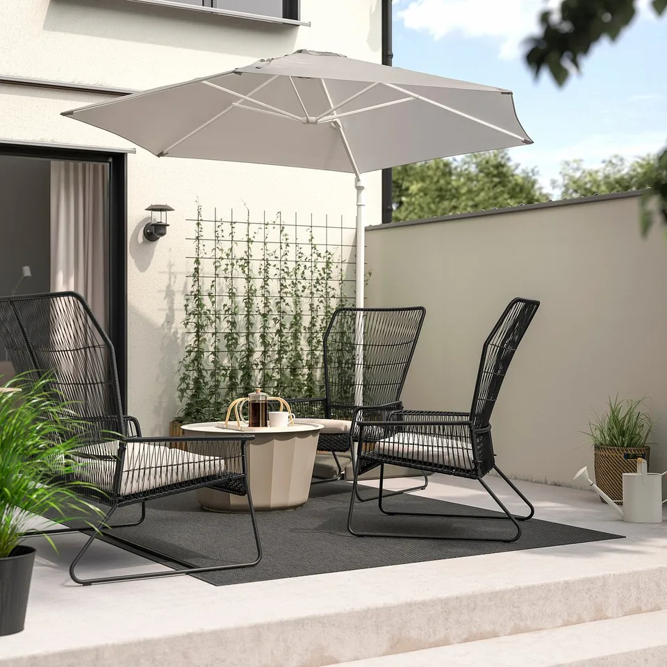 TALLSKÄR black wicker patio set under large beige umbrella on concrete balcony with white walls.