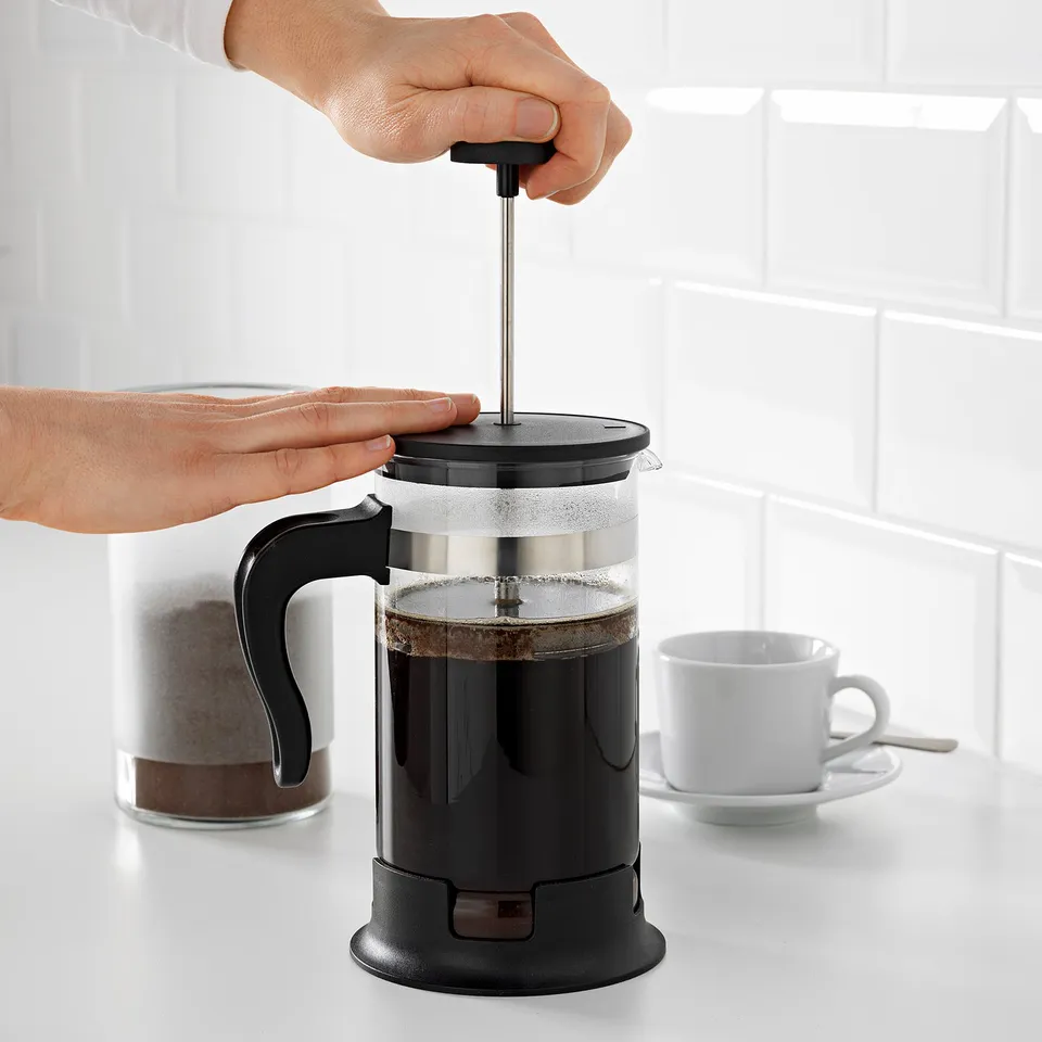 Person pressing UPPHETTA coffee maker, cup and coffee grounds nearby.