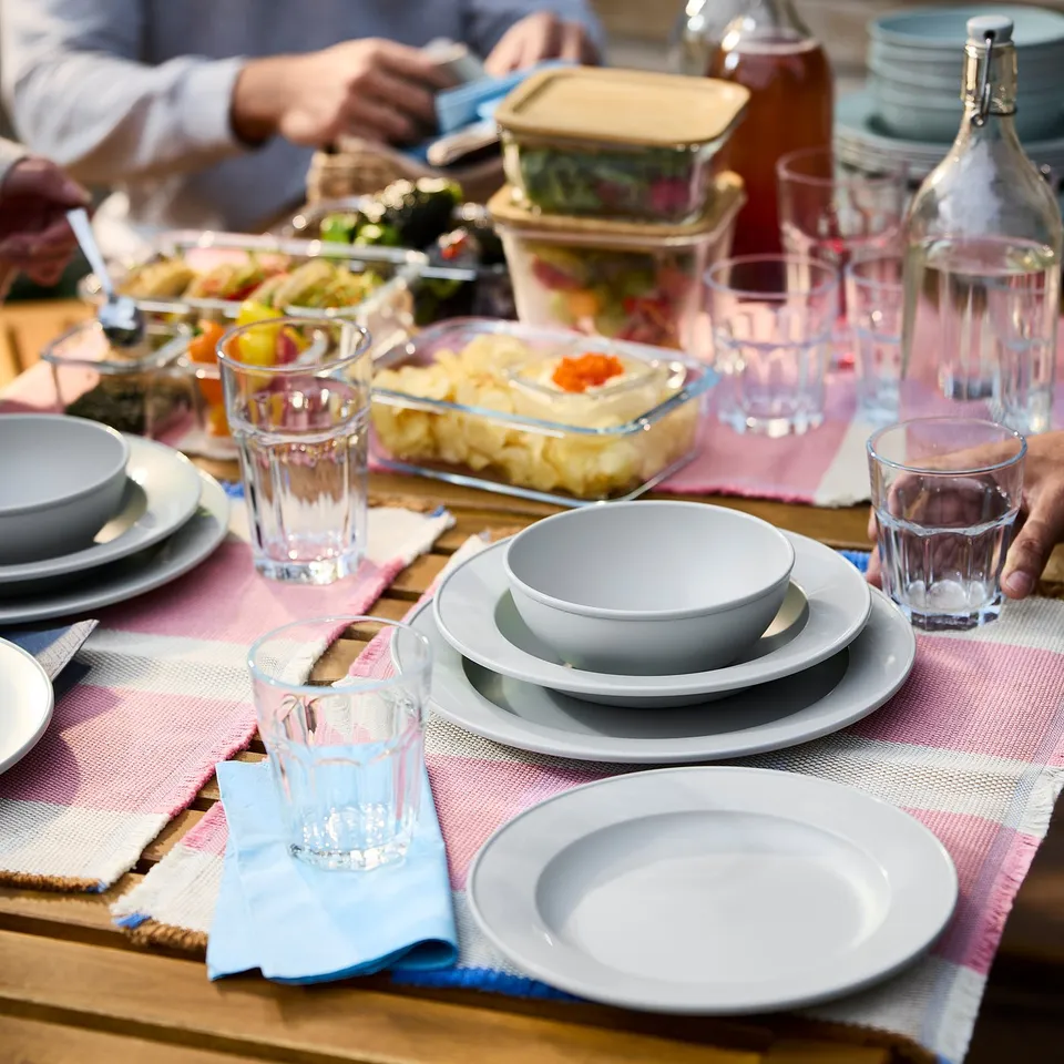 Wooden table with SIMKRABBA green-grey dinnerware, glasses, and colourful food.