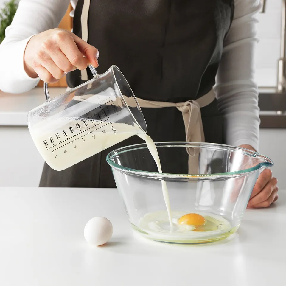 Person pours milk from a measuring jug into a bowl with an egg. The jug has markings for measuring liquid.