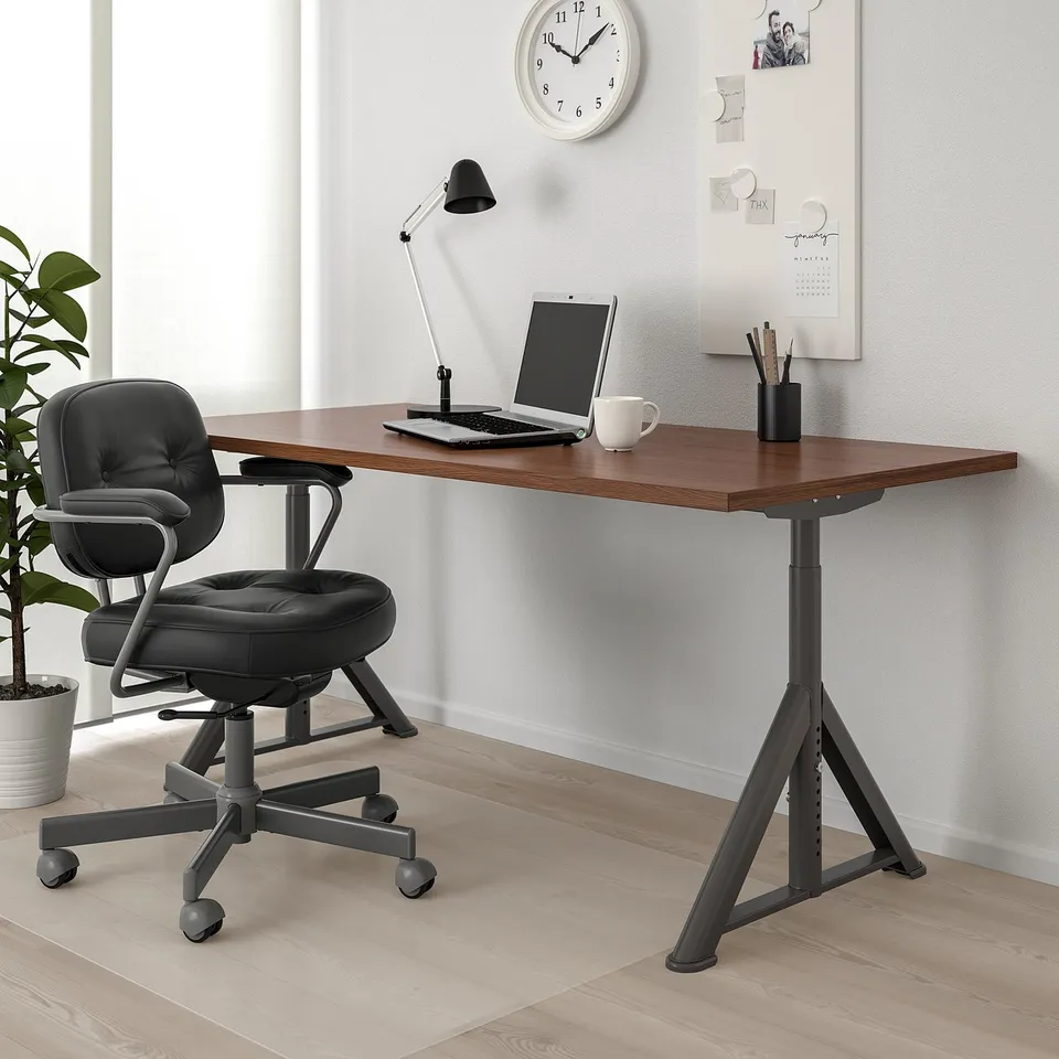 IDÅSEN desk with dark top, black legs. Laptop, mug, lamp on it. Black swivel chair. Wall clock, corkboard.