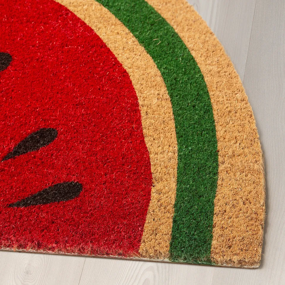 Bright red doormat with black seeds design, bordered by green and beige stripes, laid on light wooden floor.