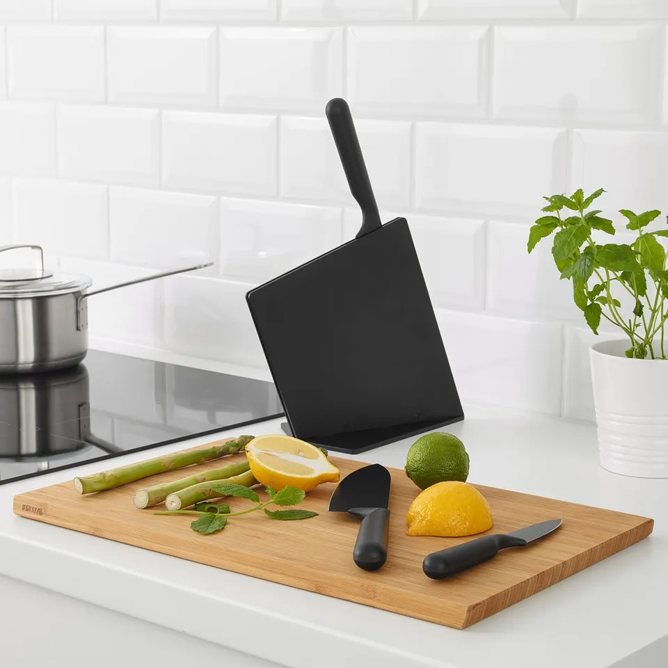 Black knife block with three knives and cutting board on white countertop. Lemons, limes, and asparagus on the board.