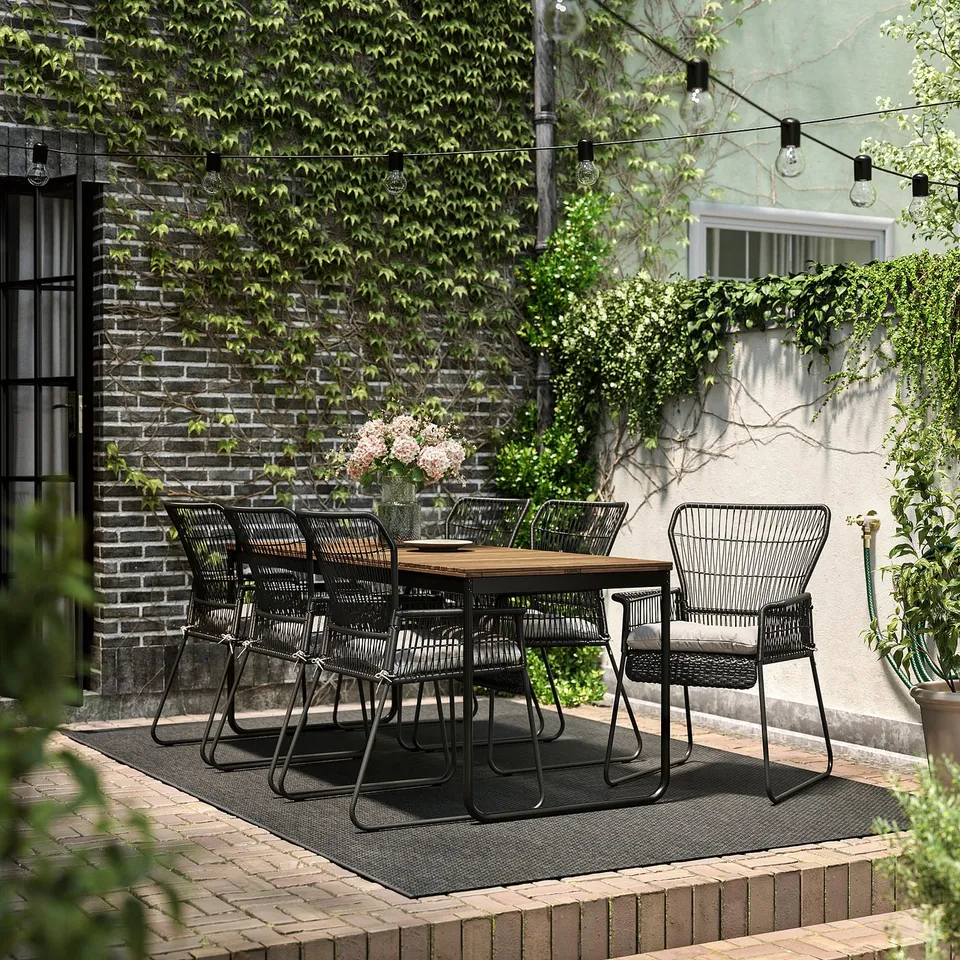 Cosy patio setup with black VÄSMAN extendable table and chairs, brick wall background, string lights.