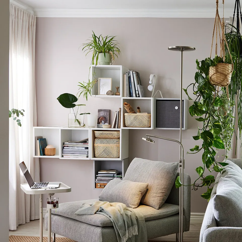 Cosy living room with EKET shelves holding books and plants. Grey chaise with pillows, green blanket, laptop table, and floor lamp.