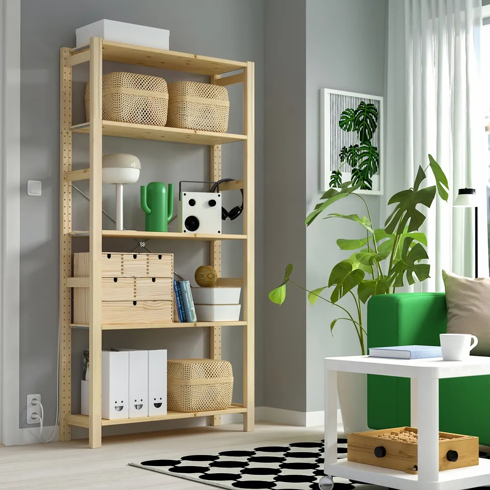 Wooden IVAR unit holds baskets, books, and a lamp, next to a large green plant against a grey wall.