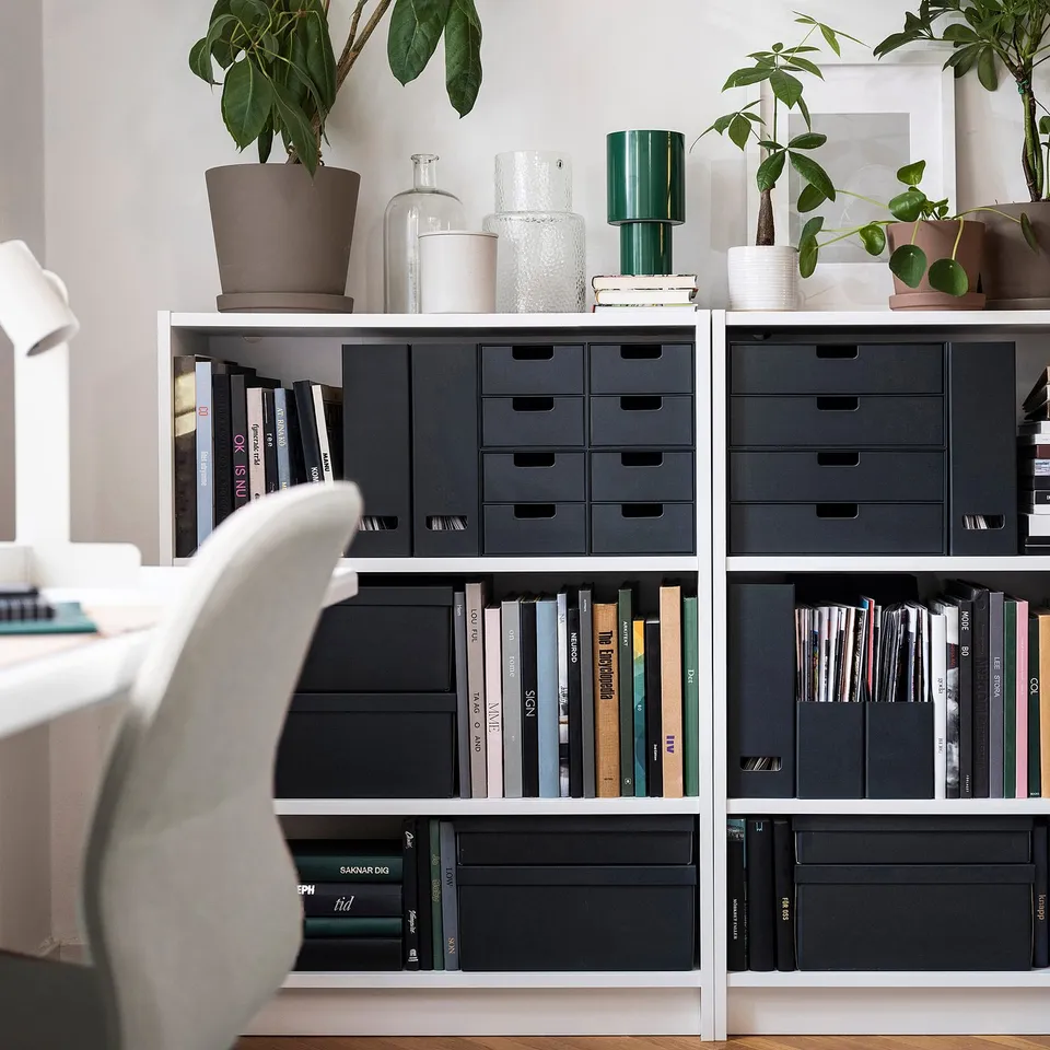 Modern bookshelf with black magazine files, books, plants, and vases. Chair partially visible.