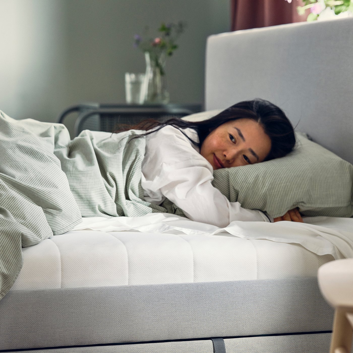 Person resting on white foam mattress in bedroom. Laptop, flowers, and glass on bedside table nearby.