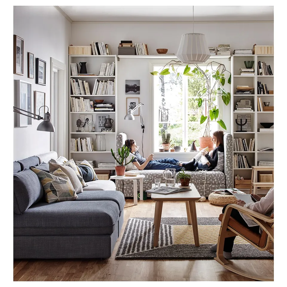 Modern living room with BILLY bookcases, family relaxing on couch, striped rug, potted plants by large window.