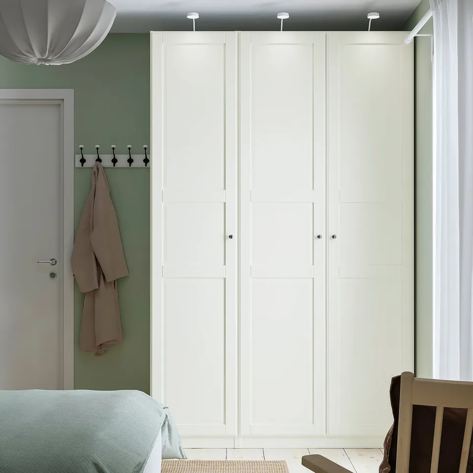 Minimalist bedroom with PAX wardrobe, light green walls. Wardrobe tall, white, fits near door. Clothes rail visible.