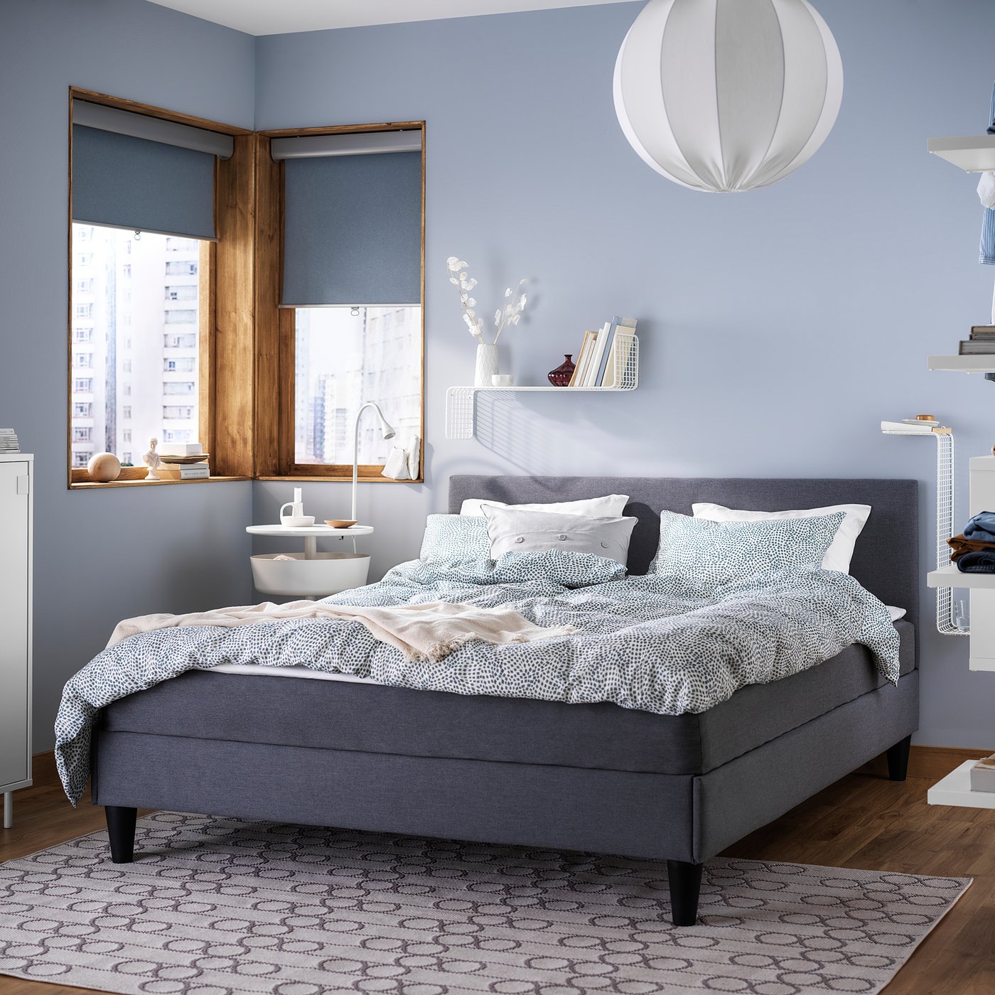 Modern bedroom with grey SÄBÖVIK bed, blue walls, white pendant light, wooden floor, and city view window.