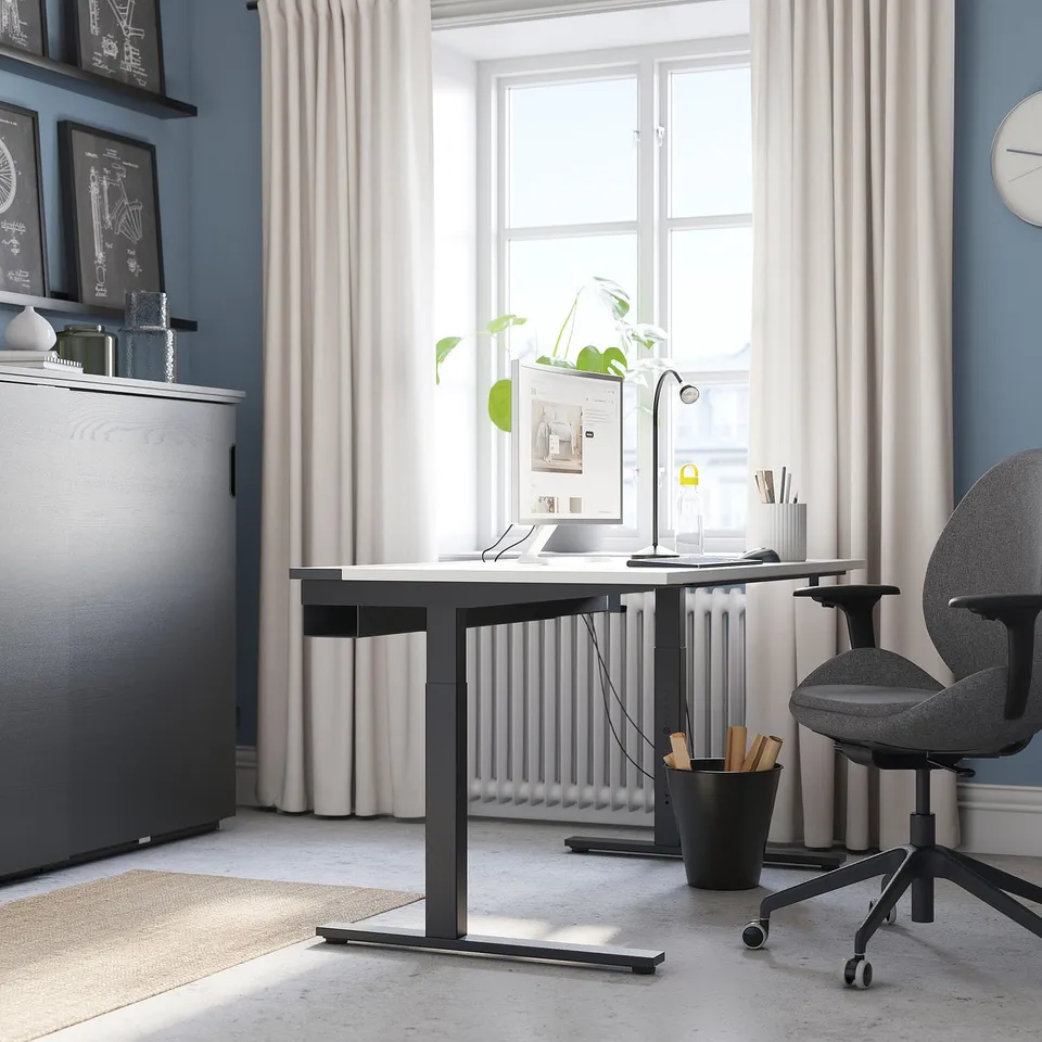 Small modern home office with a white desk, grey chair, and plants, set near a large window with sheer curtains.