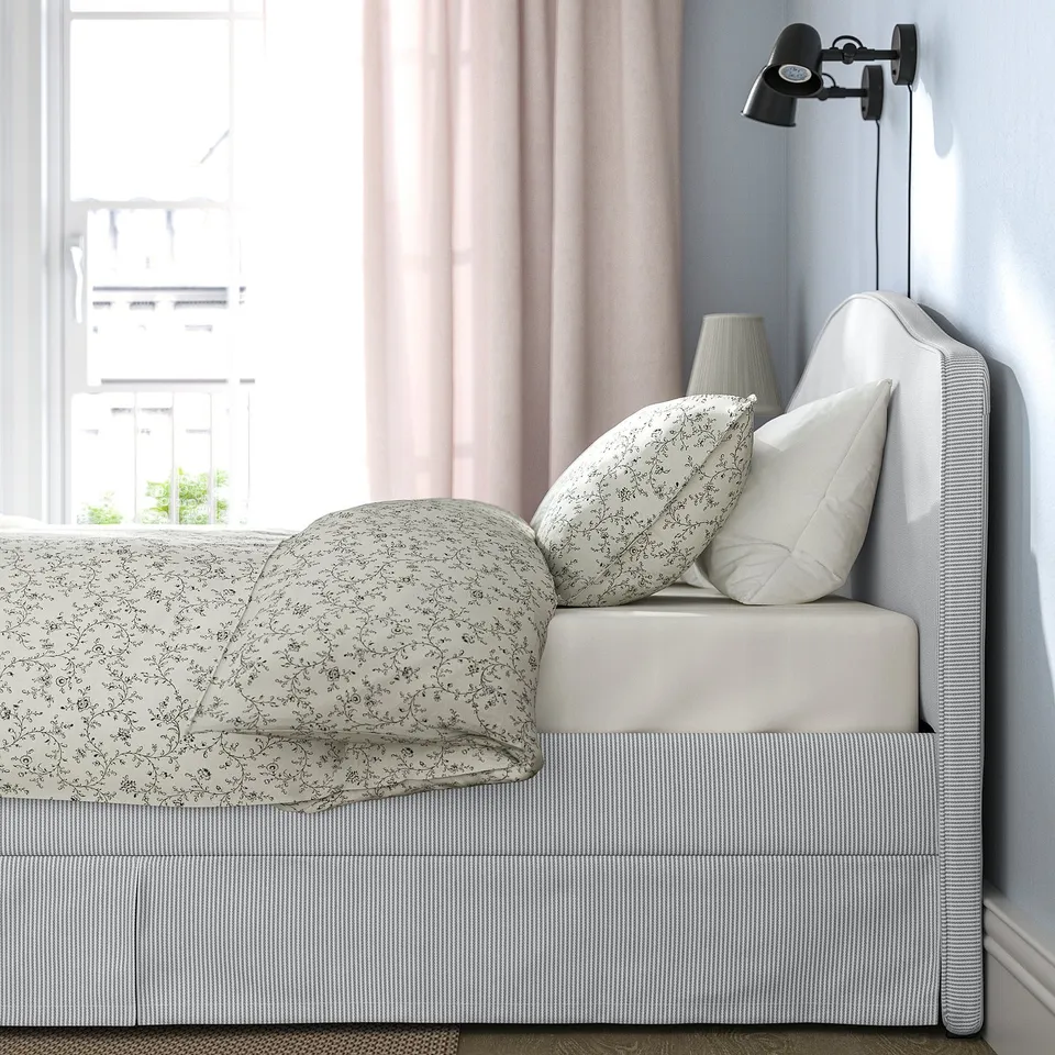 Bed with grey-white striped cover and floral patterned blanket. Includes decorative pillows and wall-mounted reading lamp.