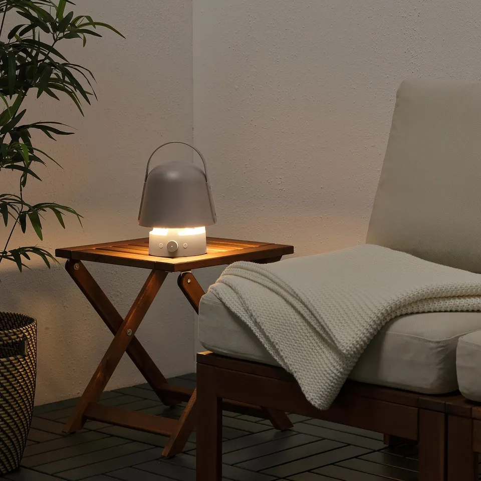 Portable lamp speaker on wooden table beside chair, bright light, cosy setup.
